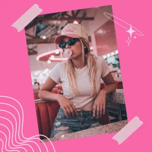Woman wearing a tan and pink “Just Out Here Vibin’” hat with black sunglasses, blowing bubblegum in a retro diner setting.