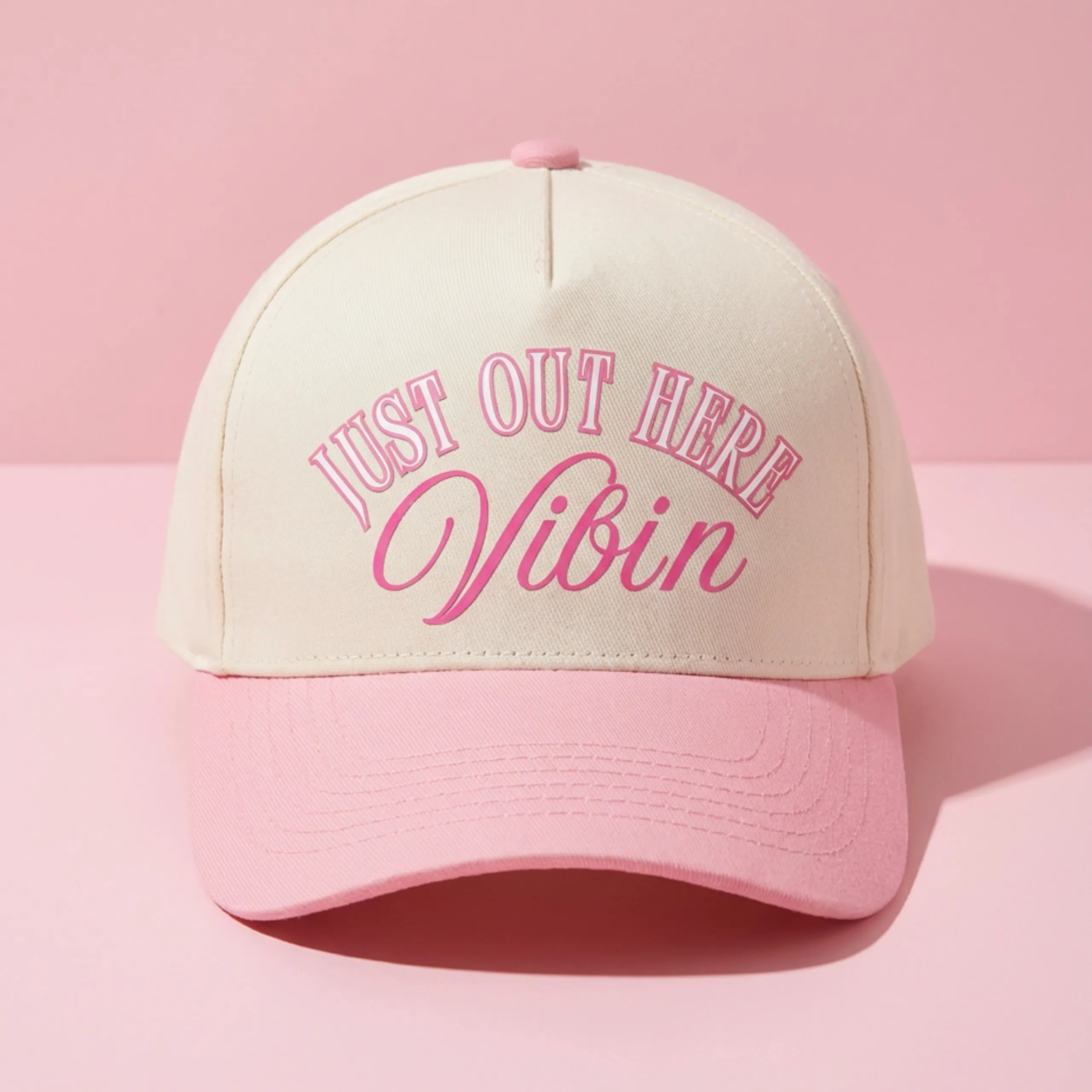 Tan and pink “Just Out Here Vibin” trucker style hat with curved pink brim on a pink background.